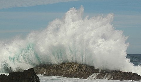 An ocean wave crashes against shoreside rocks.  Photo by Amada44. 
